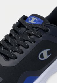 Svart sportssko med texturerad överdel, blå accenter och kontrasterande vit sula. Har platta snören och logodetalj på sidan.