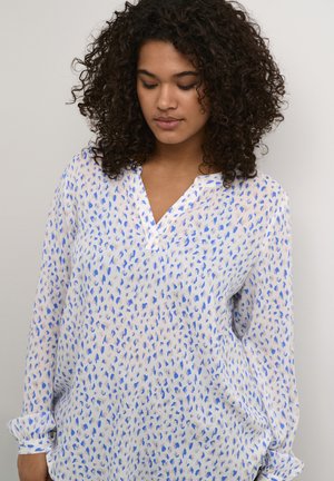 Blusa bianca con motivi blu e beige sparsi, maniche lunghe e scollo a V. Realizzata in un tessuto leggero e trasparente.