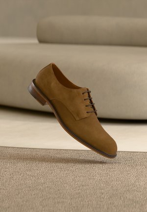 Chaussure habillée homme en cuir suédé marron avec lacets flottant au-dessus d'un tapis beige avec un canapé en arrière-plan.