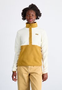 Polarowy sweter z kremową górną częścią i musztardowym dolnym panelem. Posiada wysoki kołnierz, zapięcie na guziki oraz małe logo na lewej piersi.