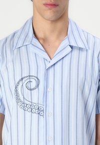 Camicia leggera azzurro chiaro a maniche corte con righe verticali bianche e blu scuro. Presenta un disegno di un polpo ricamato sul lato sinistro.