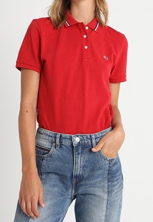 Persoon draagt een rood polo-shirt met korte mouwen en witte knopen, ingestopt in een hooggetailleerde blauwe spijkerbroek, met één hand in de zak.