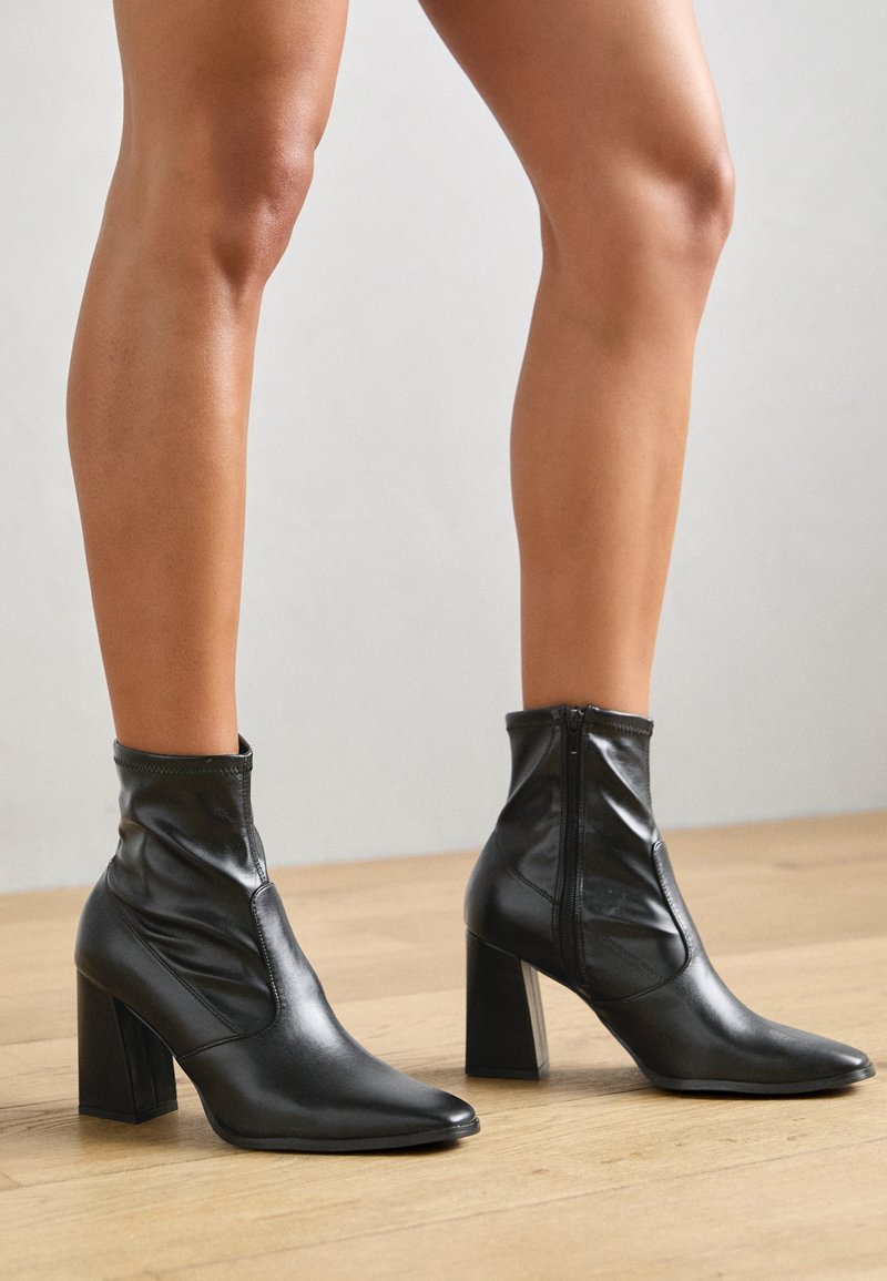 Jambes portant des bottines en cuir noir à talons carrés, debout sur un sol en bois clair contre un mur uni.