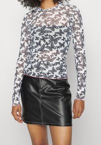 Kvinna klädd i en genomskinlig långärmad topp med svarta och grå fjärilstryck och en svart läder minikjol med vit sömnad.