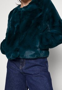 Veste en fausse fourrure teal avec un col rond, texture douce et deux poches latérales, associée à un jean en denim bleu foncé.