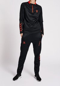 Conjunto atlético negro con top de manga larga que presenta un cierre de cuarto y acentos rojos, combinado con pantalones a juego y patrón de chevron en las mangas.