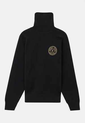 Črna polo majica iz rebraste tkanine z izrazitim zlatim krožnim logotipom na levi strani prsnega koša ter dolgimi rokavi.