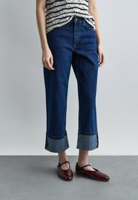 Άτομο με μανσετωμένα σκούρα μπλε τζιν, ριγέ αμάνικη μπλούζα και σκούρα κόκκινα παπούτσια Mary Jane, στέκεται σε γκρι δάπεδο.