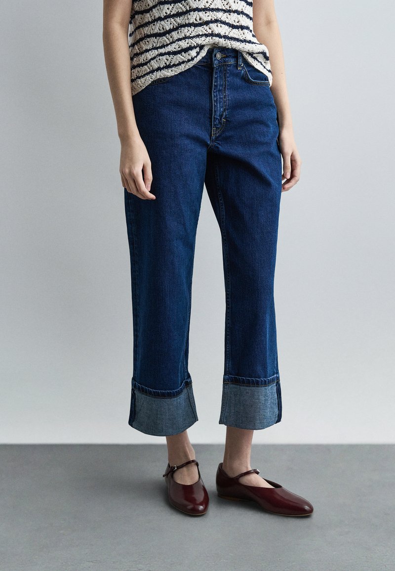 Άτομο με μανσετωμένα σκούρα μπλε τζιν, ριγέ αμάνικη μπλούζα και σκούρα κόκκινα παπούτσια Mary Jane, στέκεται σε γκρι δάπεδο.