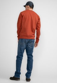 Oranje sweatshirt met geribbelde manchetten en zoom, gecombineerd met blauwe jeans en donkere schoenen. Aanzicht van achteren, met een subtiele logo-detail nabij de nek.