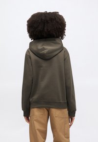 Olivovo zelená mikina s kapucňou na šnúrku, rebrovanými manžetami a lemom. Nosená s khaki cargo nohavicami, ktoré zvýrazňujú uvoľnený strih a mäkkú textúru.
