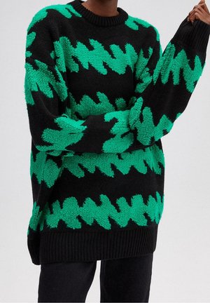 Pull noir oversize avec des motifs en zigzag dentelés vert vif porté par une personne aux bras croisés et main levée.