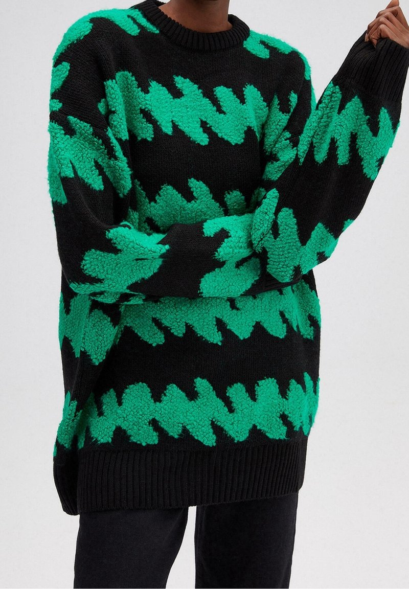Pull noir oversize avec des motifs en zigzag dentelés vert vif porté par une personne aux bras croisés et main levée.