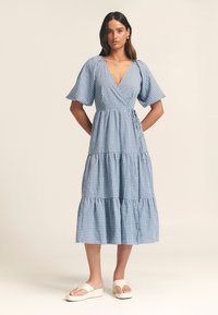 Sininen ruudullinen mekko, jossa syvä V-kaula-aukko, liehuvat lyhyet hihat, kerroksellinen helma ja vyötärönauha. Yhdistetty valkoisiin korokepohjasandaaleihin.