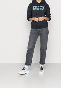 Zwarte hoodie met een blauw Levi's-logo, gecombineerd met opgerolde donkergrijze jeans en witte sneakers met blauwe accenten.
