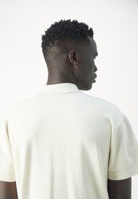 Camisa polo color crema con una textura suave, de manga corta y con cuello doblado, vista desde la parte trasera. Diseño simple, sin patrones visibles.
