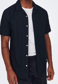 Camicia a maniche corte con bottoni di colore blu scuro, tessuto leggero, colletto aperto e bottoni bianchi. Indossata sopra una maglietta bianca.