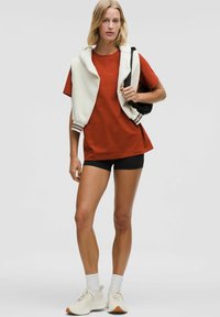 Sarkans kokvilnas t-krekls, melni šorti, krēmkrāsas hoodie pār pleciem, balti sporta apavi ar teksturētām akcentiem, sportiskais stils.