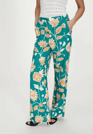 Femme portant un pantalon large couleur turquoise avec de grands motifs floraux orange et blanc, assorti à des talons ouverts noirs et un top blanc sans manches.