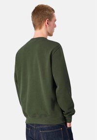 Felpa verde oliva, girocollo, polsini e orlo a costine, texture morbida, design minimalista, indossata con jeans di denim scuro.
