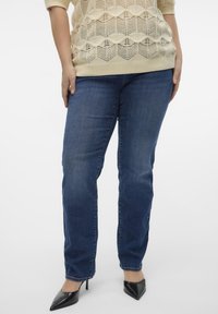 Donkerblauwe jeans in een recht model met minimale vervaging, gecombineerd met een lichtbeige gebreid topje en zwarte puntige hakken.
