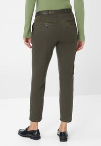 Olivengrüne Slim-Fit-Hosen mit schwarzem Bund, hinteren Taschen mit Knöpfen und geraden Säumen, kombiniert mit schwarzen Slippern.