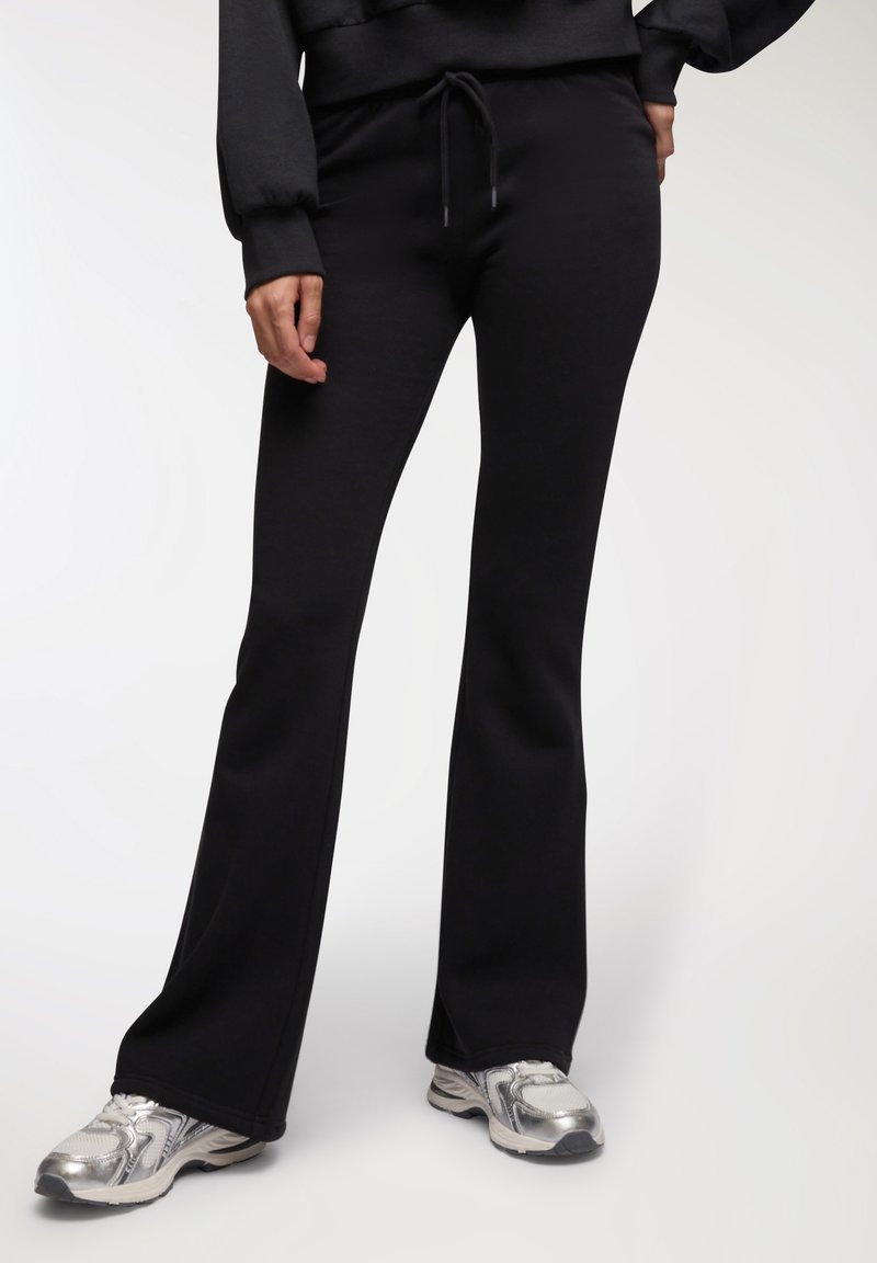 Czarne spodnie dresowe z szeroką nogawką, elastycznym pasem i sznurkiem, wykonane z miękkiego materiału, w zestawie z srebrnymi butami sportowymi.