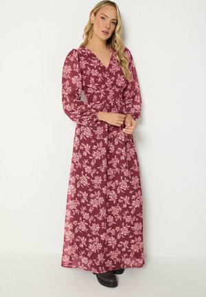 Donna che indossa un abito lungo a maniche lunghe, bordeaux con fantasia floreale e scollo a V, in piedi davanti a uno sfondo bianco neutro.