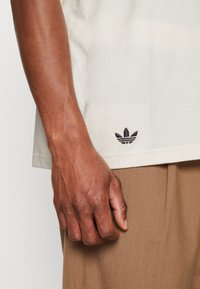 Krämfärgad bomullst-shirt med en liten svart adidas-logotyp nära den nedre fållen, med en smal passform och korta ärmar.