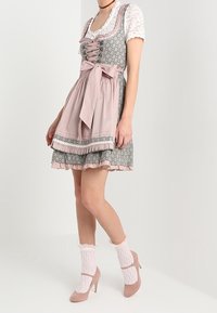 Robe dirndl traditionnelle avec une jupe à motifs rose et gris, des dentelles et un corsage ajusté avec un nœud. Accompagnée de chaussettes en dentelle blanches et de chaussures à talons.