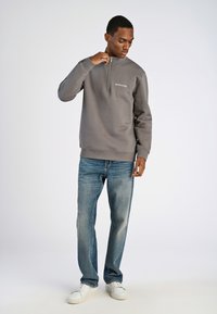 Szary sweatshirt z golfem na zamek błyskawiczny, z prążkowanymi ściągaczami przy rękawach i na dole, w zestawieniu z jasnoniebieskimi dżinsami i białymi trampkami, noszony przez osobę stojącą.