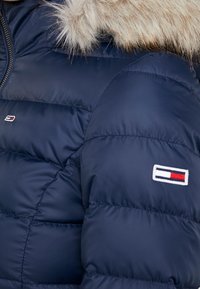 Granatowa pikowana kurtka puchowa z kapturem wykończonym sztucznym futrem. Posiada przeszycia na sekcjach oraz małą naszywkę z logo w kolorach czerwonym, białym i niebieskim.