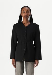 Veste noire avec un col en V, des manches longues, un fermeture à boutons et une ceinture. Fabriquée en tissu au toucher lisse et dotée de deux poches avant.