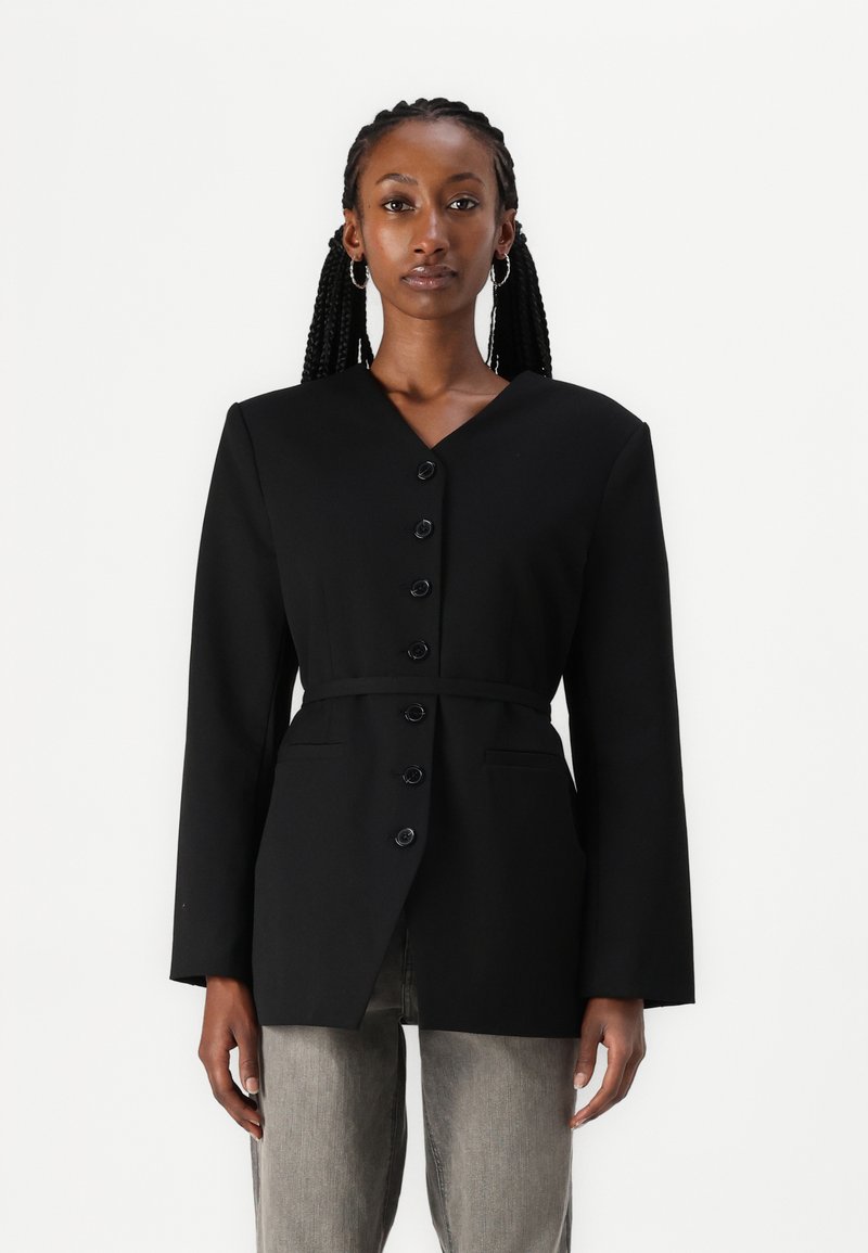 Veste noire avec un col en V, des manches longues, un fermeture à boutons et une ceinture. Fabriquée en tissu au toucher lisse et dotée de deux poches avant.