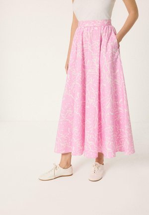 Vrouw draagt een lange rok met hoge taille, roze bloemenprint en zakken, een wit mouwloos top en crèmekleurige platte veterschoenen.