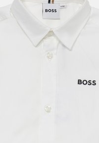 Balta marškiniai su klasikine apykakle, kuriuose yra juodas išsiuvinėtas "BOSS" logotipas ir skaidrūs mygtukai priekyje.