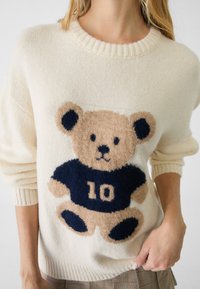 Kvinna som bär en krämfärgad tröja med ett nallebjörnsdesign, en mörkblå skjorta och siffran 10 på framsidan.