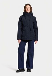 Marinblå vattentät jacka med huva, två framfickor och dragkedja. Matchas med vida jeans och svarta stövlar.