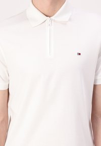 Witte poloshirt van gladde katoen, met een halve rits aan de voorkant en een klein geborduurd logo in rood, wit en blauw.
