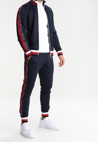 Marineblauwe trainingspak met rode en witte gestreepte accenten, ritsjack en elastische broekspijpen, gecombineerd met witte sneakers.