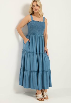 Frau trägt ein blaues, ärmelloses, gestuftes Maxikleid mit Taschen und silbernen, gewebten Pantoffeln, steht vor einer schlichten, hellen Wand.