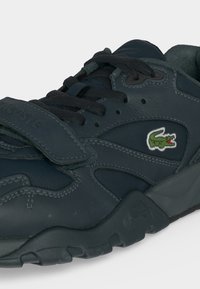 Σκούρο ναυτικό μπλε αθλητικό παπούτσι Lacoste με μαύρα κορδόνια, λουρί βέλκρο και το πράσινο λογότυπο του κροκοδείλου στο πλαϊνό πάνελ.