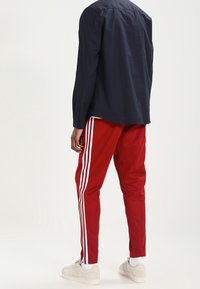 Červené sportovní kalhoty s bílými bočními pruhy, doplněné tmavě modrou košilí na knoflíky a béžovými teniskami. Na botách je viditelné logo.