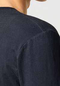 Acercamiento a la parte superior de la espalda y el hombro de una persona que lleva una camiseta de manga larga de color azul marino oscuro, de canalé, con costuras visibles.