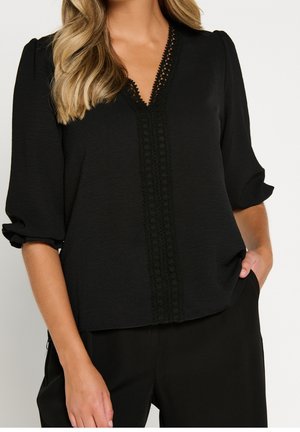 Femme portant un chemisier noir avec un col en V bordé de dentelle et des manches bouffantes 3/4, associé à un pantalon noir, main dans la poche.