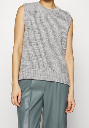 Pull sans manches en tricot gris porté avec un pantalon en cuir vert sarcelle taille haute, montrant le torse et les bras sur un fond uni.