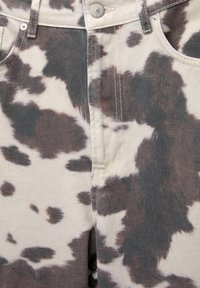 Gros plan sur un bouton et une braguette sur un pantalon beige avec un motif imprimé vache noir et marron.