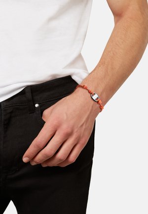Poignet masculin avec un bracelet en cordon orange noué comportant une petite fermeture métallique, main reposant près de la poche d’un pantalon noir et ourlet de chemise blanche visible.