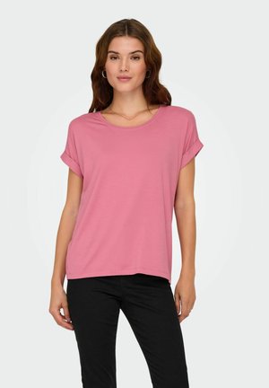 Donna con t-shirt rosa a maniche corte con risvolto e pantaloni neri, in piedi contro uno sfondo chiaro e semplice.