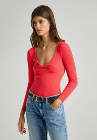 Maglietta rossa a maniche lunghe con righe, scollatura a V profonda e dettaglio al centro con nodo, abbinata a jeans azzurri a vita alta e una cintura nera.